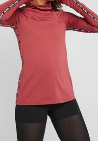 Rotes sportliches Langarmoberteil mit hohem Kragen und schwarzen Seiteneinsätzen, auf denen der Text "NIKE PRO" steht. Hergestellt aus leichtem, dehnbarem Material.