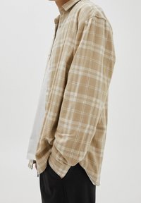 Persona vistiendo una camisa de cuadros beige y blanca abotonada sobre una camiseta blanca, con las manos en los bolsillos de un pantalón negro sobre un fondo liso.