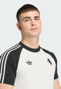 Κοντομάνικη μπλούζα με κρέμ κορμό και μαύρα ραγκλάν μανίκια, με υφασμένους λογότυπους και λευκές ρίγες στα μανίκια.