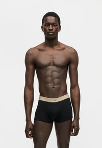 Ένας μυώδης άνδρας στέκεται στραμμένος μπροστά φορώντας μαύρα ανδρικά εσώρουχα Tommy Hilfiger με μπεζ μέση, σε ένα απλό λευκό φόντο.