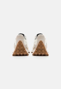 Zapatillas deportivas con parte superior de ante beige, acentos blancos y suela de goma texturizada. Presenta un distintivo talón triangular con agarres circulares.