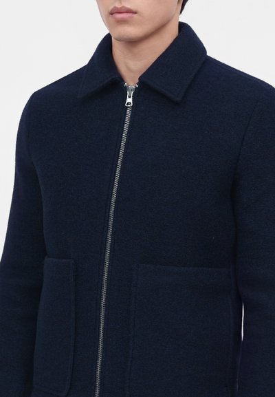 Homme portant une veste en laine bleu marine foncé zippée avec col et deux grandes poches avant, visible du cou à la taille.