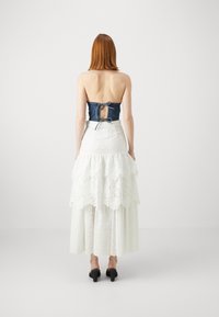 Top sin tirantes de mezclilla con un detalle de lazo en la parte posterior, combinado con una falda maxi blanca de encaje en capas. Presenta texturas superpuestas y colores contrastantes.