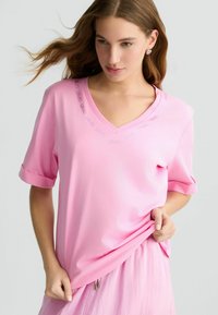 T-shirt a V in rosa chiaro realizzato in tessuto morbido, con maniche corte arrotolate e un logo discreto attorno al collo. Texture liscia, vestibilità comoda.