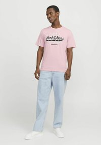 Rosa Kurzarm-T-Shirt mit schwarzem "Jack & Jones" Grafiktext. Getragen mit hellblauen Schlagjeans und weißen Sneakers.