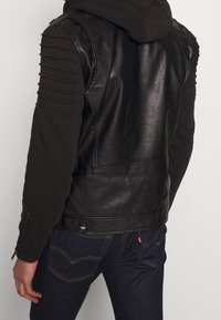 Giacca in pelle nera con dettagli a coste sulle maniche, cappuccio nero incorporato e tasche con zip. Presenta un design aderente e una texture liscia.
