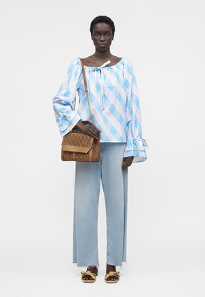 Γυναίκα φοράει ανοιχτό μπλε καρό μπλούζα, τζιν ξεθωριασμένο με φαρδιά πόδια, ταμπά υφαντό τσαντάκι ώμου και ταμπά σανδάλια με λεπτομέρεια φιόγκο, στέκεται μπροστά σε λευκό φόντο.