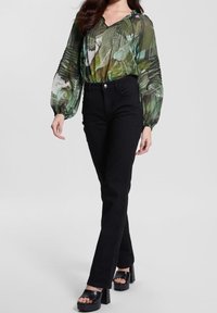 Nainen, jolla on yllään läpinäkyvä vihreä pikkulehtikuvioinen paita, mustat korkeavyötäröiset housut ja mustat korkeat kantapäät, seisoo tasaisella taustalla.