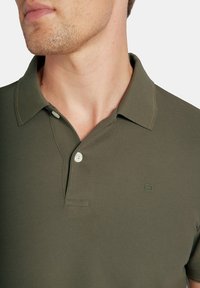 Polo w kolorze oliwkowym, wykonany z teksturowanej tkaniny, z klasycznym kołnierzykiem, dwoma guzikami i małym logo na klatce piersiowej.