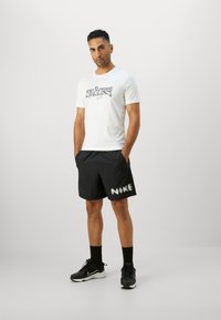 Camisa blanca con estampado gráfico negro y logotipo, combinada con pantalones cortos negros que tienen el texto "NIKE" en blanco. Modelo llevando zapatillas y calcetines negros.
