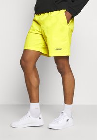 Žluté kraťasy s elastickým pasem, malým černým logem Adidas, spárované s bílými teniskami a ponožkami. Krátká, lehká tkanina.