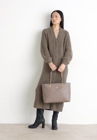 Brun uld lang cardigan, høj talje med knapper, båret over en tætsiddende beige top. Sorte knæhøje støvler og en kviltet grå håndtaske med kædestropper.