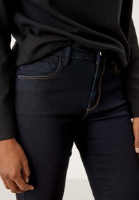 Jean en denim bleu foncé à coupe slim. Il présente des coutures orange et cinq poches, avec un bouton en métal à la taille.