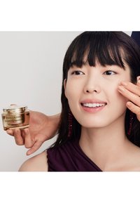 Estee Lauder Revitalizing Supreme+ kräm i en klar glasburk med ett guldlock, hålls av en hand nära en leende kvinna med mörkt hår.