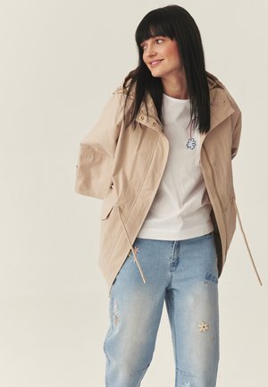 Donna sorridente con capelli neri e lisci indossa una giacca con cappuccio beige sopra una camicia bianca e jeans chiari azzurri ricamati, su sfondo neutro.