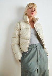 Doudoune beige avec un col haut, fermeture éclair à l'avant et texture matelassée. Associée à un pantalon gris avec taille élastique et poches latérales.