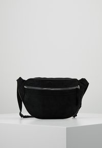 Borsetta in suede nera con una tasca con cerniera silver e tracolla regolabile, poggiata su una superficie bianca contro uno sfondo grigio uniforme.