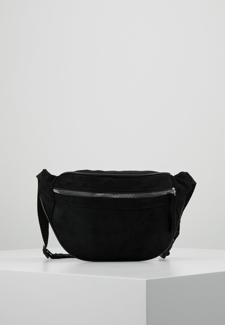 Borsetta in suede nera con una tasca con cerniera silver e tracolla regolabile, poggiata su una superficie bianca contro uno sfondo grigio uniforme.