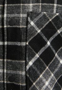 Svart och vit rutigt flanelltyg med ett diamantmönster, med svarta knappar och en synlig ficka på vänster sida.