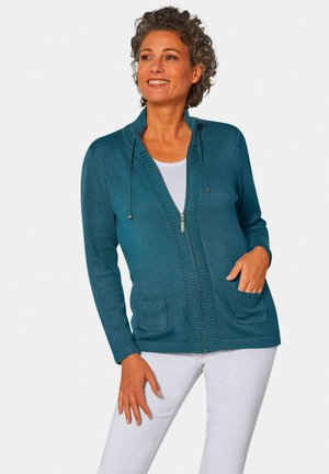 Cardigan teal avec une fermeture éclair sur le devant, une capuche et deux poches avant. Présente des bords côtelés et un tissu tricoté texturé. Porté par-dessus un haut blanc.