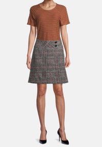 Hnědý texturovaný top se zvlněným designem, doplněný sukní s kostkovaným vzorem sahající po kolena, která má černé a růžové akcenty. Vzhled dotvářejí černé vysoké podpatky.