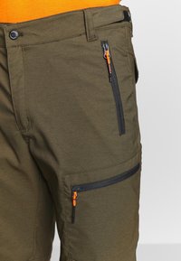 Oliivinvihreät shortsit mustilla vetoketjuilla ja oransseilla vetoketjun vetimillä sivutaskuissa, yhdistettyinä oranssiin puseroon.