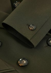 Detail tmavě zelené tkaniny s hladkým povrchem, která obsahuje dva lesklé knoflíky a manžetu s neatním zakončením.
