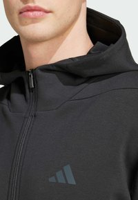 Haut de survêtement noir zippé en tissu lisse, avec un col haut et un logo gris discret sur la poitrine. Gros plan sur l'encolure et le visage.