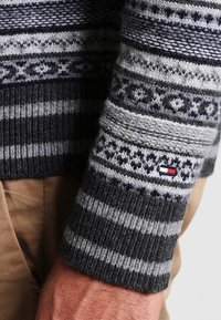 Pull en maille gris et bleu marine avec des rayures verticales et horizontales et des motifs géométriques. Petite étiquette logo sur le poignet. Texture douce.