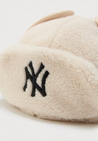 Un bonnet en polar couleur crème avec des lettres NY noires brodées qui se chevauchent à l'avant, reposant sur une surface blanche.