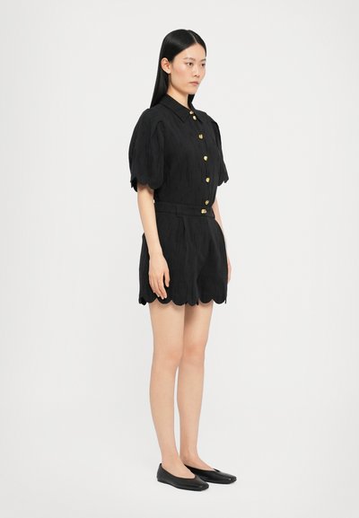 Schwarzer Kurzarm-Overall mit kragenartigem Hals, goldenen Knöpfen, gewelltem Saum und strukturiertem Stoff. Kombiniert mit schwarzen flachen Schuhen.