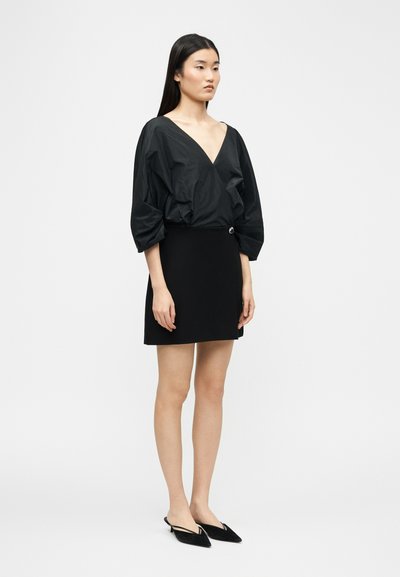 Femme debout portant un haut noir à encolure en V avec manches bouffantes, une mini-jupe noire avec détail boucle, et des ballerines noires à bout pointu et bride arrière.