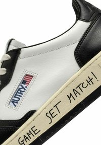 Sneaker in pelle bianca e nera con un design retrò, caratterizzati dalla scritta "GAME SET MATCH!" stampata di lato e un'etichetta con logo.