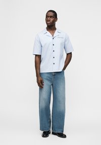 Άνδρας που φοράει γαλάζιο πουκάμισο με κοντά μανίκια και κουμπιά, φαρδιά μπλε τζιν παντελόνια και μαύρα παπούτσια, στέκεται με το ένα χέρι στην τσέπη.