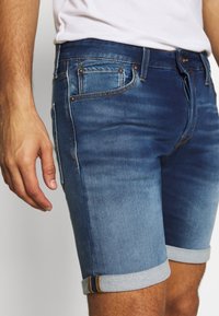 Denim šortky v středně modré barvě, s úzkým střihem, ohrnutými manžetami s kontrastním prošíváním a jemným pamětinkováním kolem kapes.