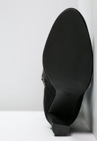 Semelle de chaussure noire avec grip texturé et petit talon, positionnée à la verticale contre une surface blanche en angle.