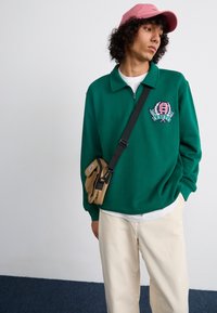 Grønn hettegenser med glidelås og brodert logo, beige skulderveske med svarte detaljer, lyse bukser og en rosa caps.