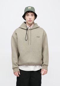 Sudadera beige con capucha, bolsillo frontal y logo negro. Lleva puesta sobre una camisa blanca. Sombrero tipo pescador gris con logo. Pantalones cortos cargo negros.