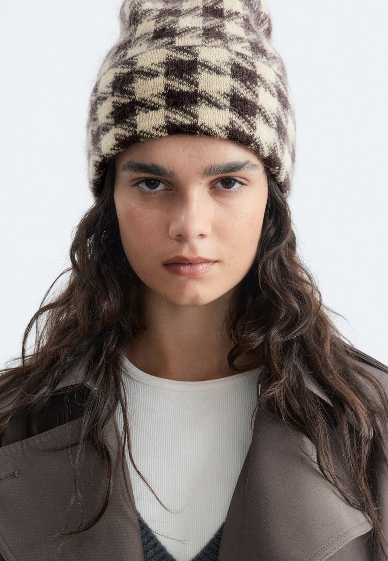 Jeune femme aux longs cheveux foncés portant un bonnet en tricot à carreaux marron et crème, un manteau marron et un haut blanc, regardant droit devant avec une expression neutre.