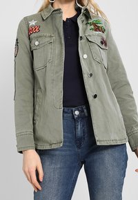 Vihreä puuvillatakki, jossa on kaksi rinnan taskua, hopeanväriset napit ja brodeeratut tarrat, mukaan lukien puu ja ananas. Klassinen kaulus ja hihansuut.