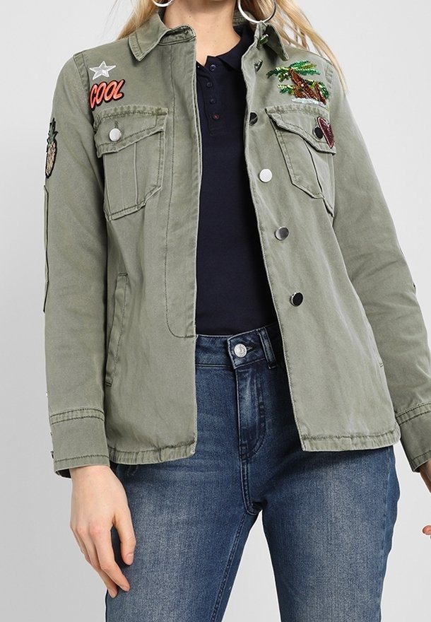 Vihreä puuvillatakki, jossa on kaksi rinnan taskua, hopeanväriset napit ja brodeeratut tarrat, mukaan lukien puu ja ananas. Klassinen kaulus ja hihansuut.