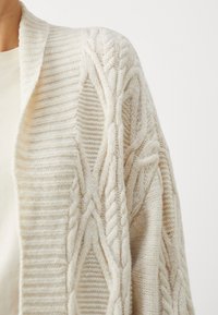 Krémově zabarvený pletený cardigan s tlustými kroucenými vzory, nošený přes krémový top s kulatým výstřihem, zobrazený od ramene po střed trupu.