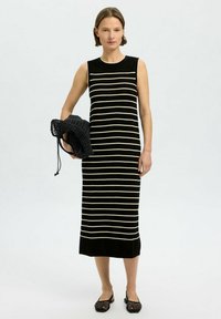 Robe sans manches rayée noir et crème avec une coupe droite, fabriquée en tissu doux, associée à un sac en tricot noir et des chaussures plates.