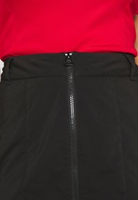 Černá sukně s předním zipem, který má lesklý kovový design. V kombinaci s červeným topem, hladká textura a přizpůsobený střih.