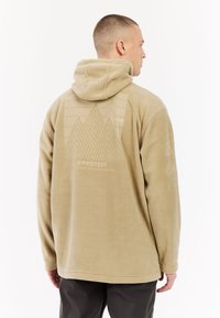 Tans kleurige fleecetruien met een gestructureerd ontwerp op de achterkant, voorzien van driehoekige patronen en branding. Zacht materiaal met een losse pasvorm.