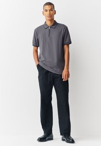 Polo met korte mouwen in donkergrijs met een gestructureerd patroon, een ritskraag en contrasterende zwarte biezen, gecombineerd met losse zwarte broek en schoenen.