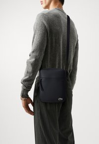 Sort læder crossbody taske har en glat overflade, lynlåset hovedrum og et lille logo detaljeret på fronten. Justerbar strop.