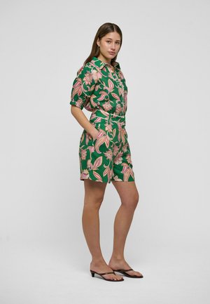 Mlada žena stoji s rukama u džepovima, nosi zelenu košulju s cvjetnim uzorkom i odgovarajuće kratke hlače te crne sandale na petu.