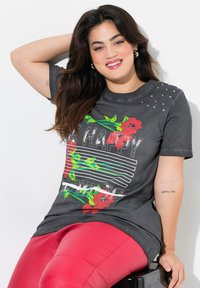 Grå grafisk t-shirt med blomsterdesign og regnbue-striber, udsmykket med sølvnitter på skulderen, parret med røde leggings.