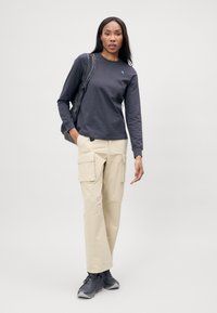 Langsleeve donkergrijze shirt met blauw logo, beige cargobroek met zijzakken en grijze sneakers, gedragen met een zwarte rugzak.